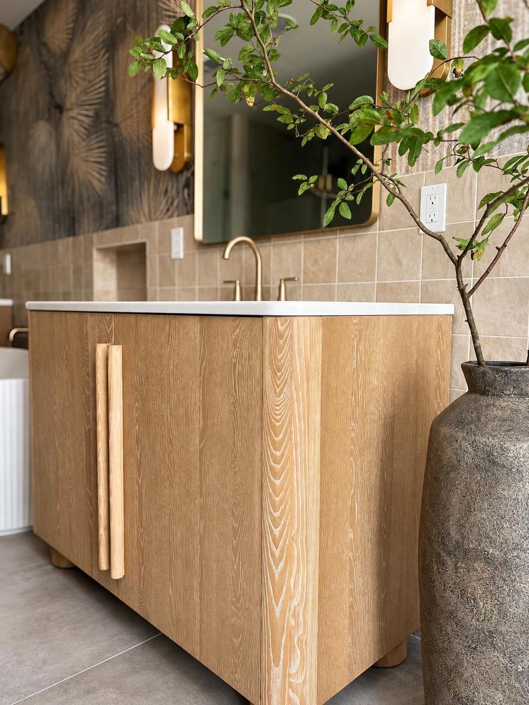 Primary bathroom vanity detail with a natural oak cabinet, brass fixtures, white quartz countertop, and a textured ceramic vase with leafy branches.