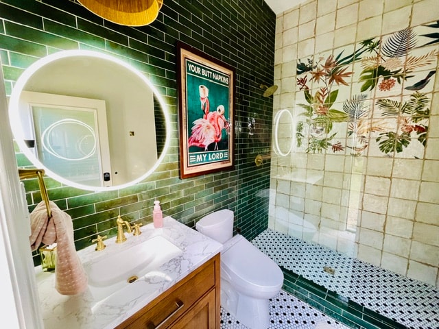 Eclectic bathroom with glossy green tile, an illuminated mirror, botanical wall tile, and brass fixtures.