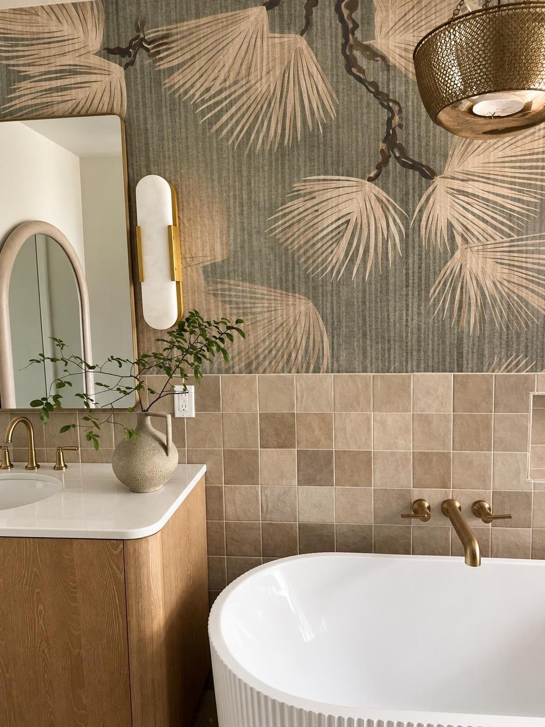 Primary bathroom vanity with an arched plaster mirror, brass sconce, ceramic vase with greenery, and a sculptural fluted tub beside warm zellige tile.